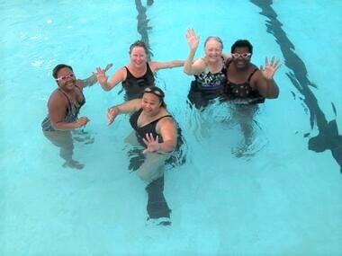 five adults in a swimming pool waving at camera